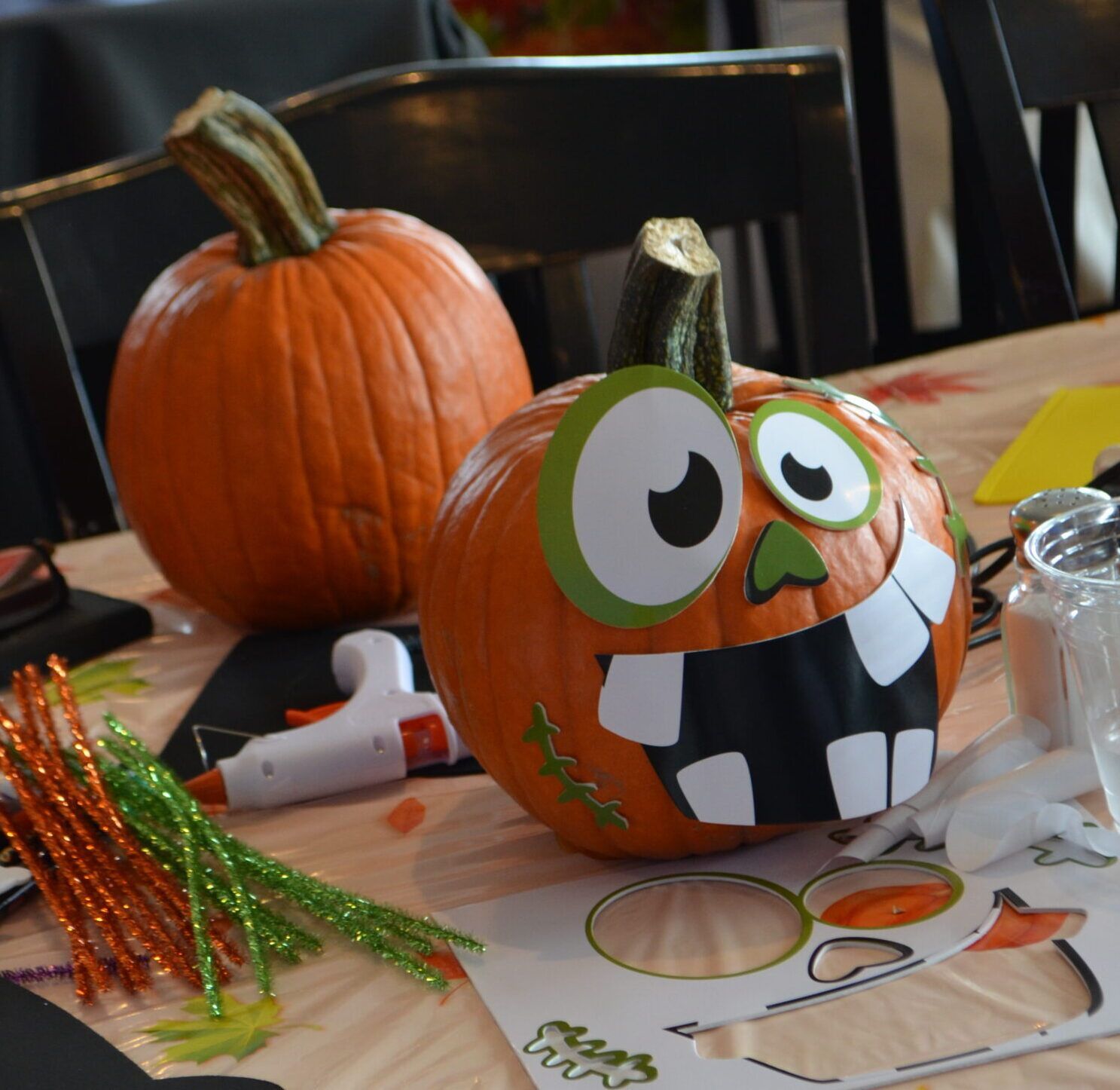 Two pumpkins on a table; one decorated as a monster with paper teeth and eye. Craft supplies scattered around.