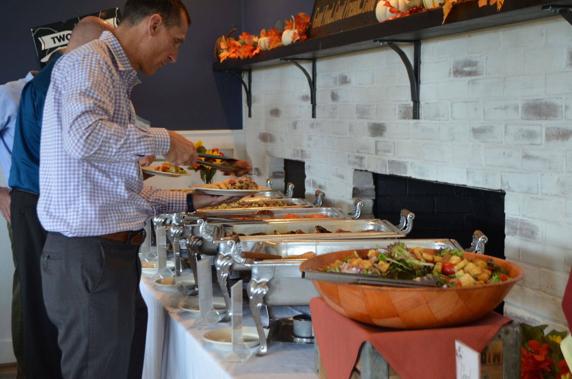 People at a buffet, serving themselves food.  The food is in warming trays on a table with a brick wall backdrop.