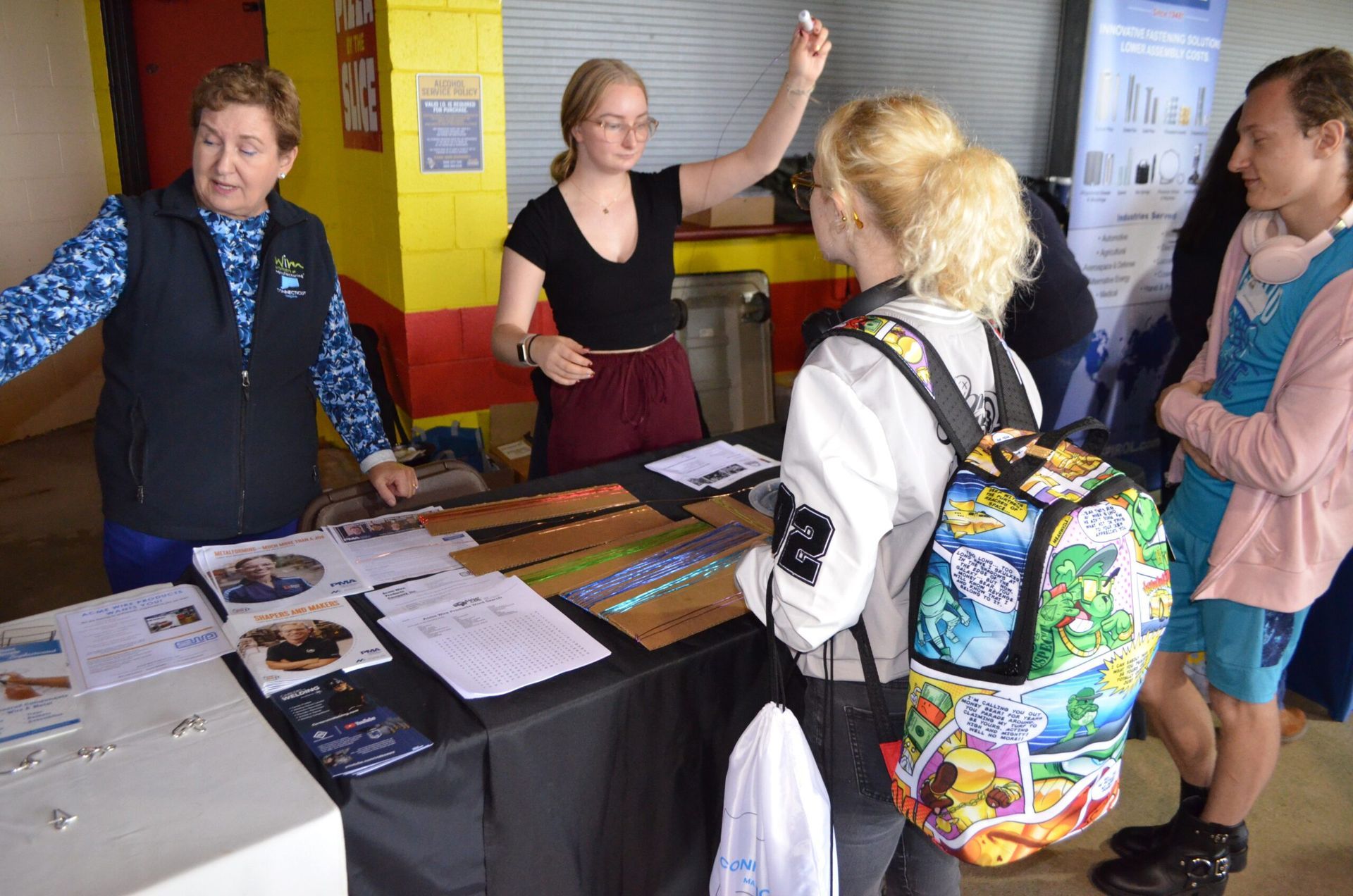 People at a display table, discussing and pointing. A woman speaks while others listen and look at materials.
