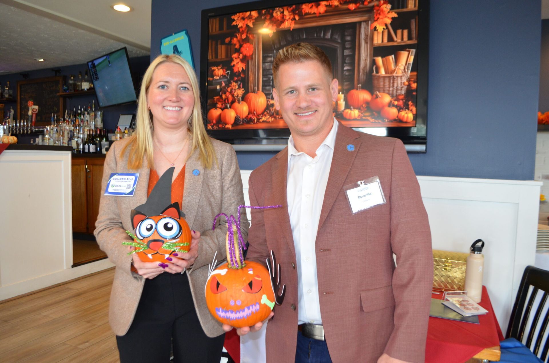 Woman and man holding painted pumpkins at an event. They are in front of a pumpkin-themed backdrop.