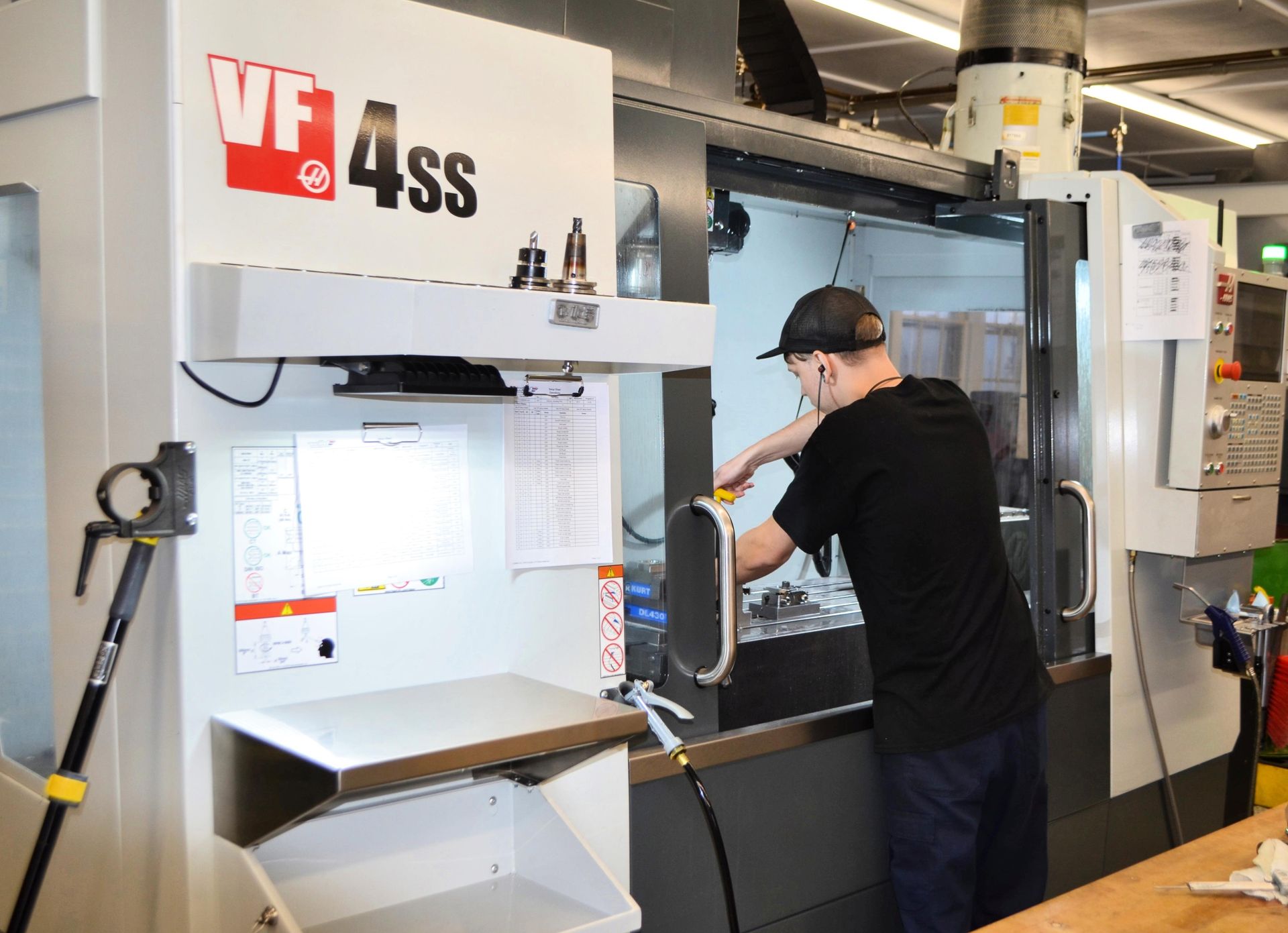 Man working on a VF4SS CNC machine in a workshop. He wears a hat and dark clothes, and is holding a tool.