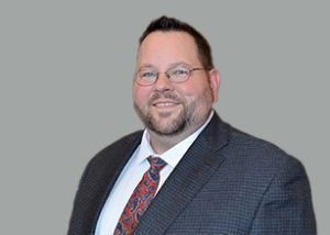 Man with glasses, wearing a suit and tie, smiles, against a gray background.