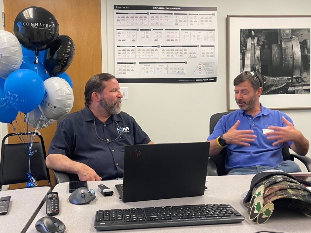 Two men sit at a table, discussing. Balloons in the background. One man gestures with his hands. A laptop and keyboard are on the table.