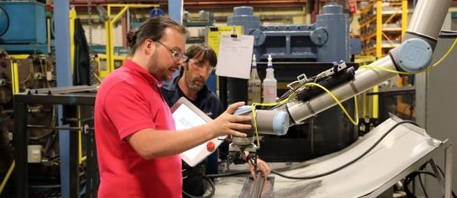 Two men work with a robotic arm in a factory setting. One man in red shirt holds a tablet.