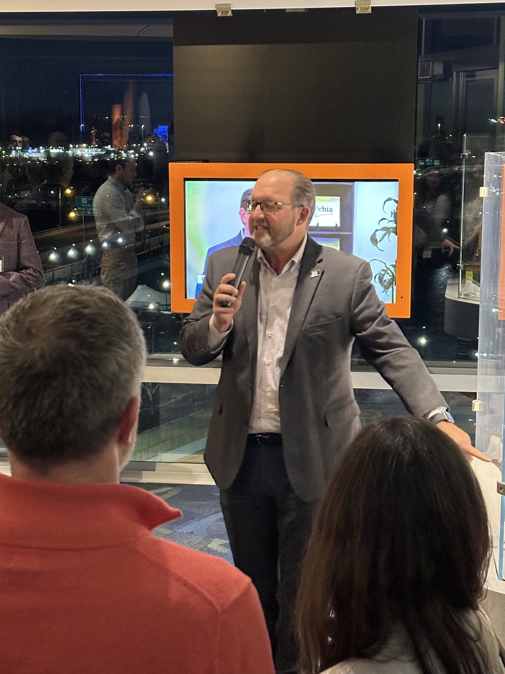 Man speaking into a microphone, gesturing, in front of a crowd, a screen, and a city view.