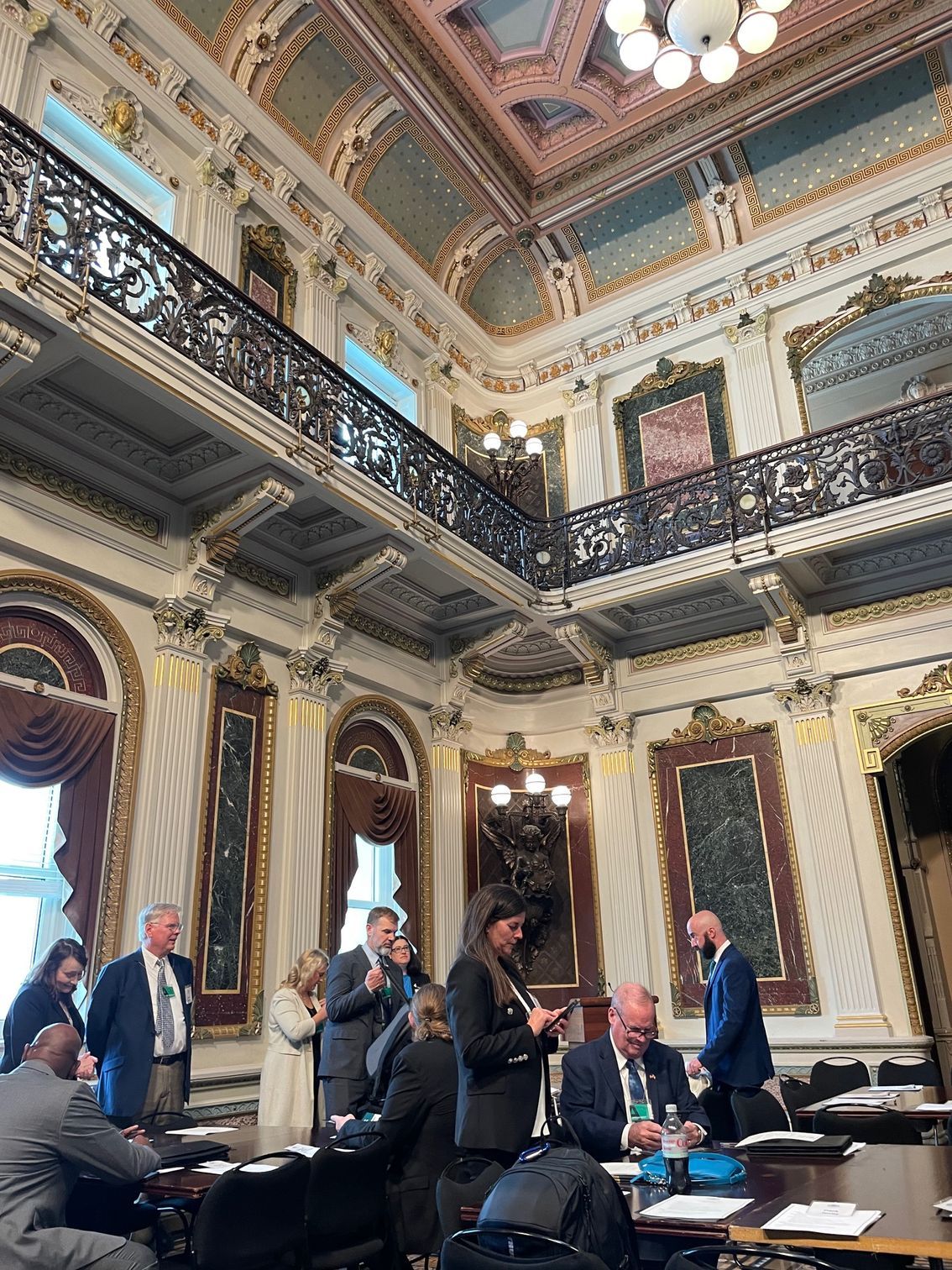 People in ornate room with high ceilings, discussing at a table. Balcony and architectural details visible.