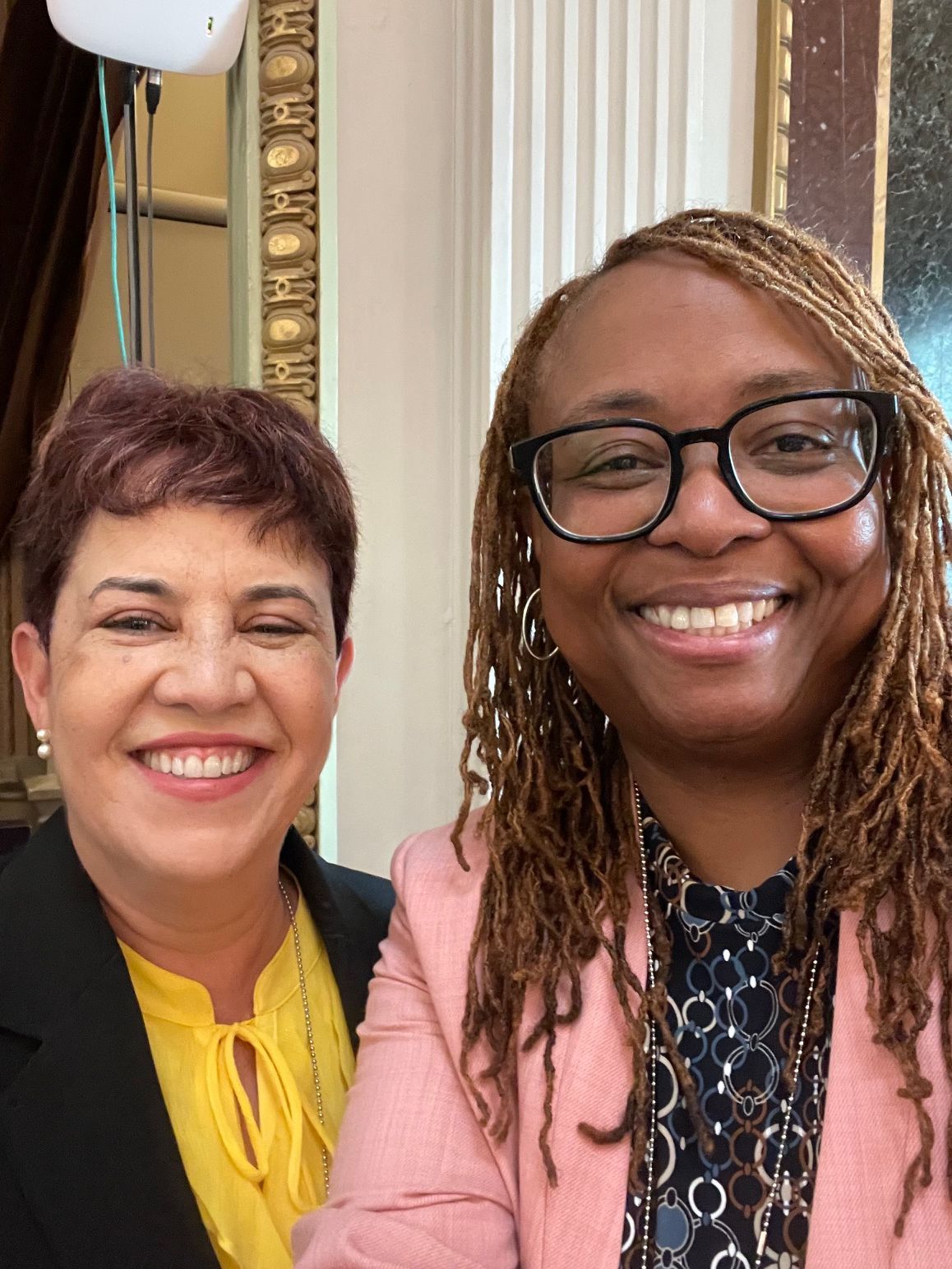 Two women smiling at camera. One wears glasses, pink jacket; other, yellow top, black jacket. Indoor setting.