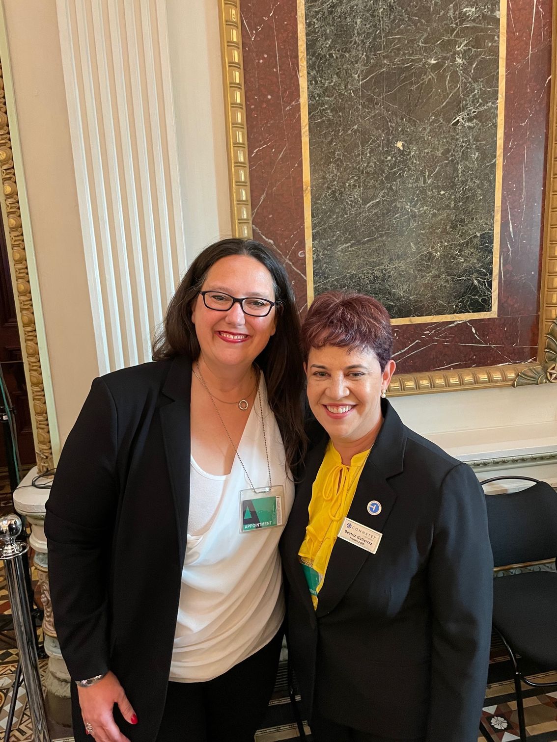 Two women standing together indoors, smiling. One wears a black blazer over a white shirt, the other a black blazer over a yellow scarf.