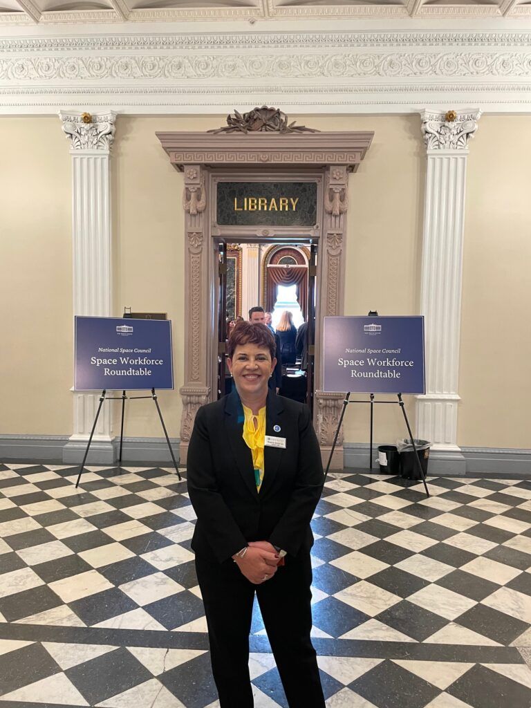 Woman in suit stands in front of a library door. A sign on the wall is partially visible. Checkered floor.