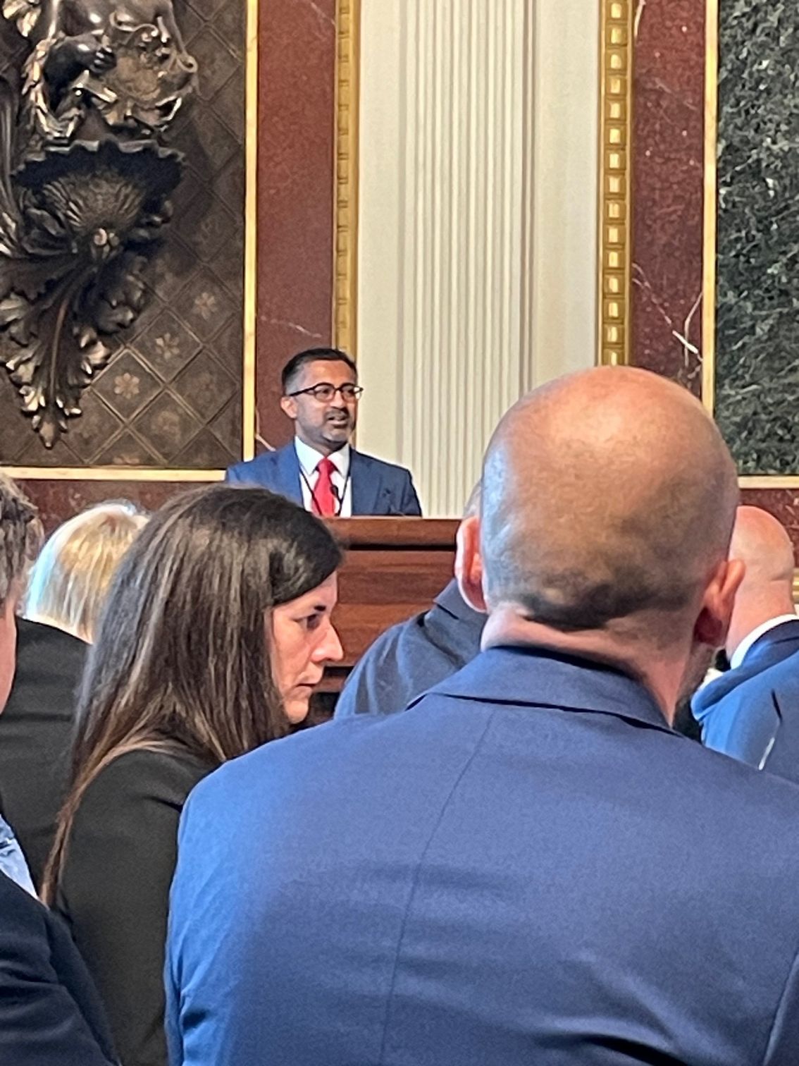 Man in suit speaks at podium, other people watch. Ornate interior.
