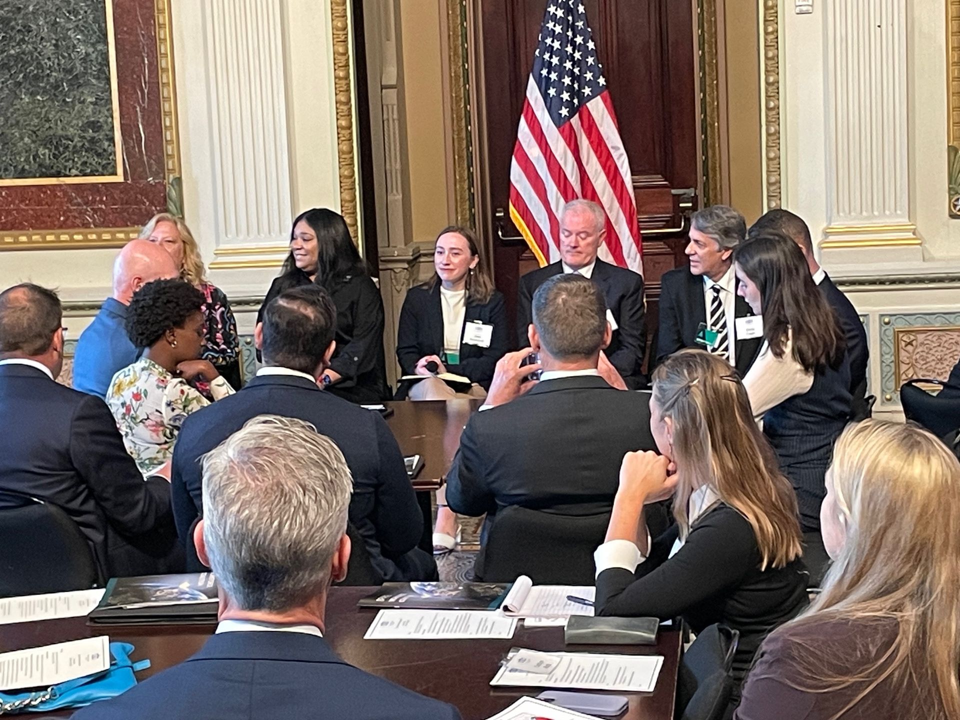 A panel discussion with people seated around a table, an American flag in the background.