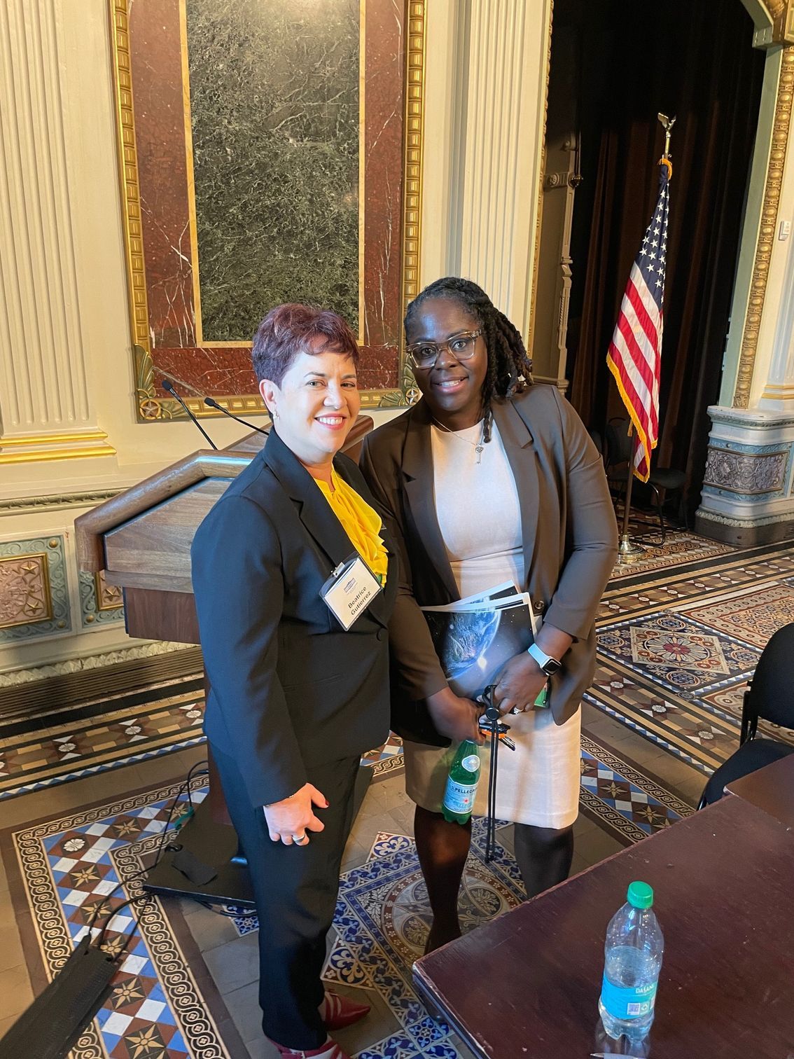 Two people smiling, posing together indoors with an American flag visible in the background.