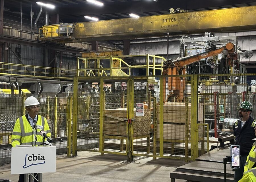 Man speaking at podium in factory, robotic arm, wood stacks, yellow safety barriers.