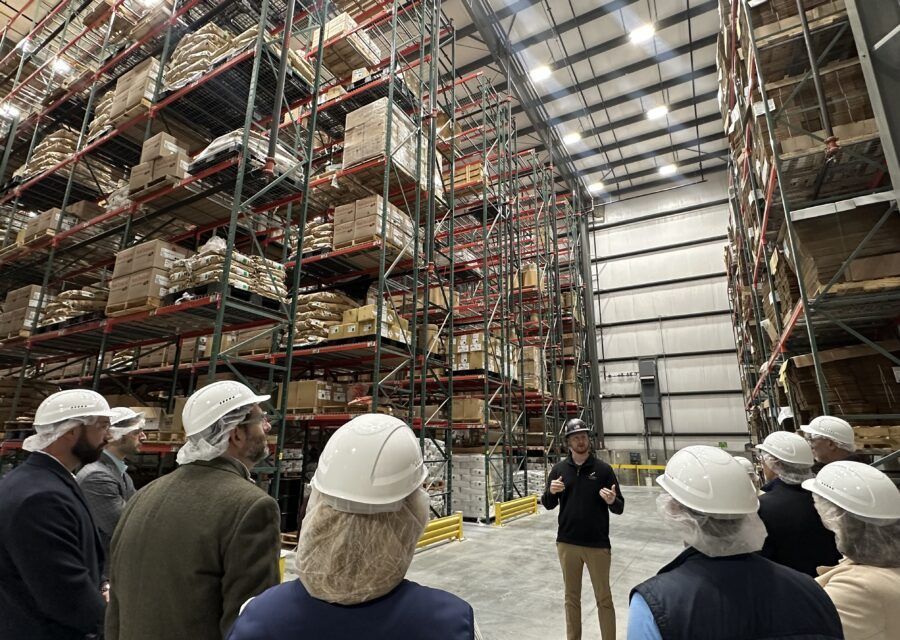 People in hairnets and hard hats listening to a man speaking in a large warehouse with shelving filled with boxes.