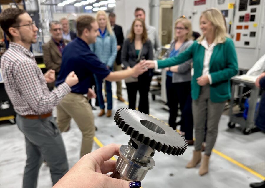 Group of people in a factory setting. A woman in green jacket greets a man as another holds a gear.