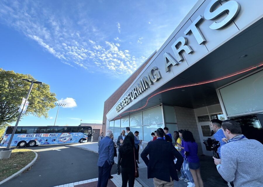 Exterior of a performing arts center with people gathered; a bus is parked nearby under a bright, blue sky.