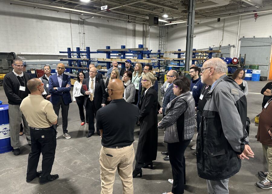Group of people in a warehouse listening to two men. Blue shelving and barrels are in the background.