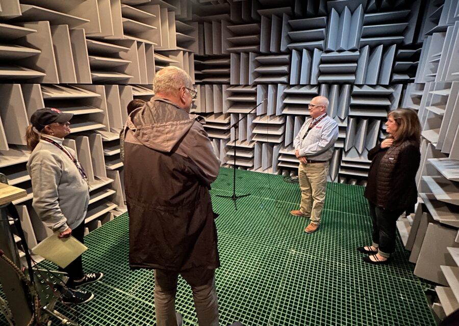 Four people in a soundproof room with noise-absorbing wedges. They are standing around a microphone.
