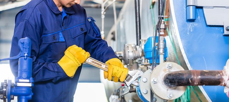 A worker wearing a blue jumpsuit and yellow gloves uses a wrench on a pipe.