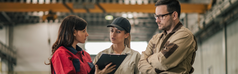 Three people in workwear discuss, holding a clipboard, indoors. Two women and a man, possibly in a factory setting.