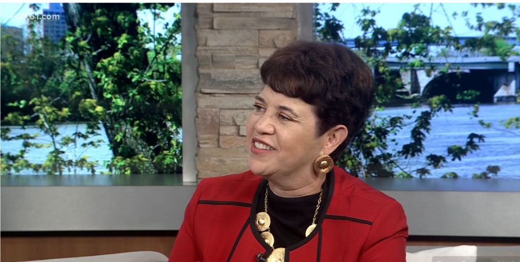 Woman in red blazer on TV set, smiling. Background: river and greenery.