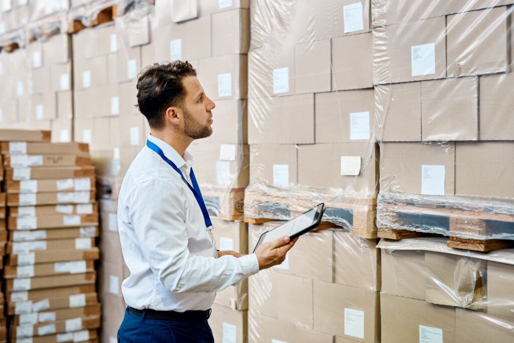 Man in white shirt and blue pants inspects stacked boxes in a warehouse, holding a tablet.