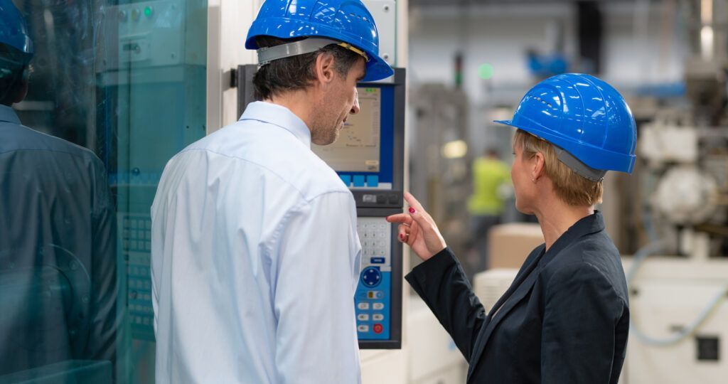 Two people in blue hard hats examining a control panel in a factory setting.