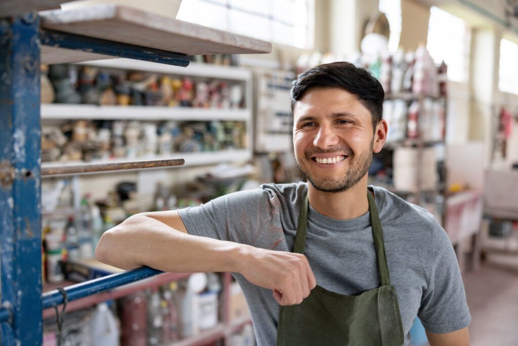 Man in workshop smiles, leans on blue support. Background: shelves with art supplies.
