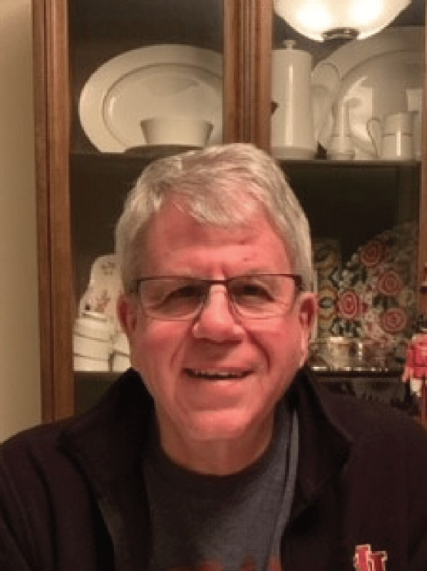A man wearing glasses is smiling in front of a glass cabinet filled with dishes.