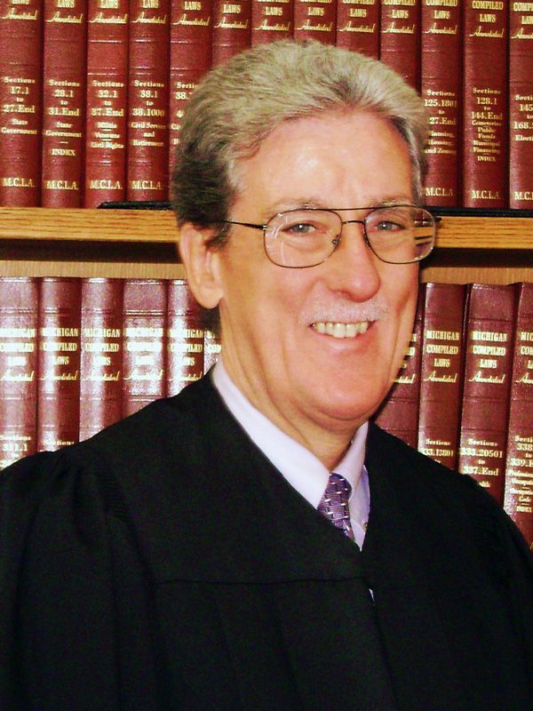 A man in a judge 's robe stands in front of a bookshelf