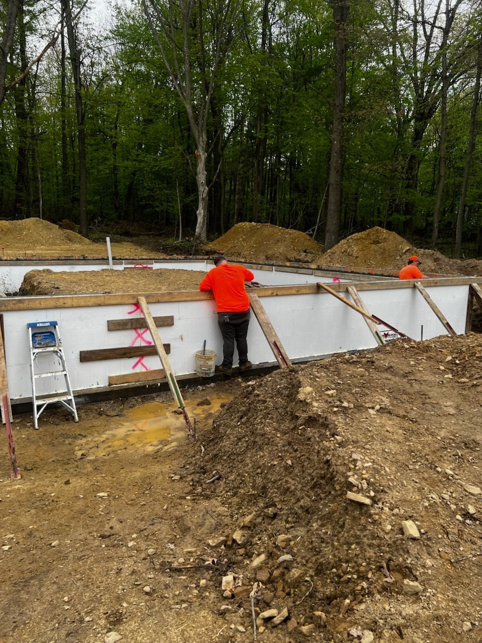 Two construction workers building a foundation in a wooded area, with a white foam structure and dirt.