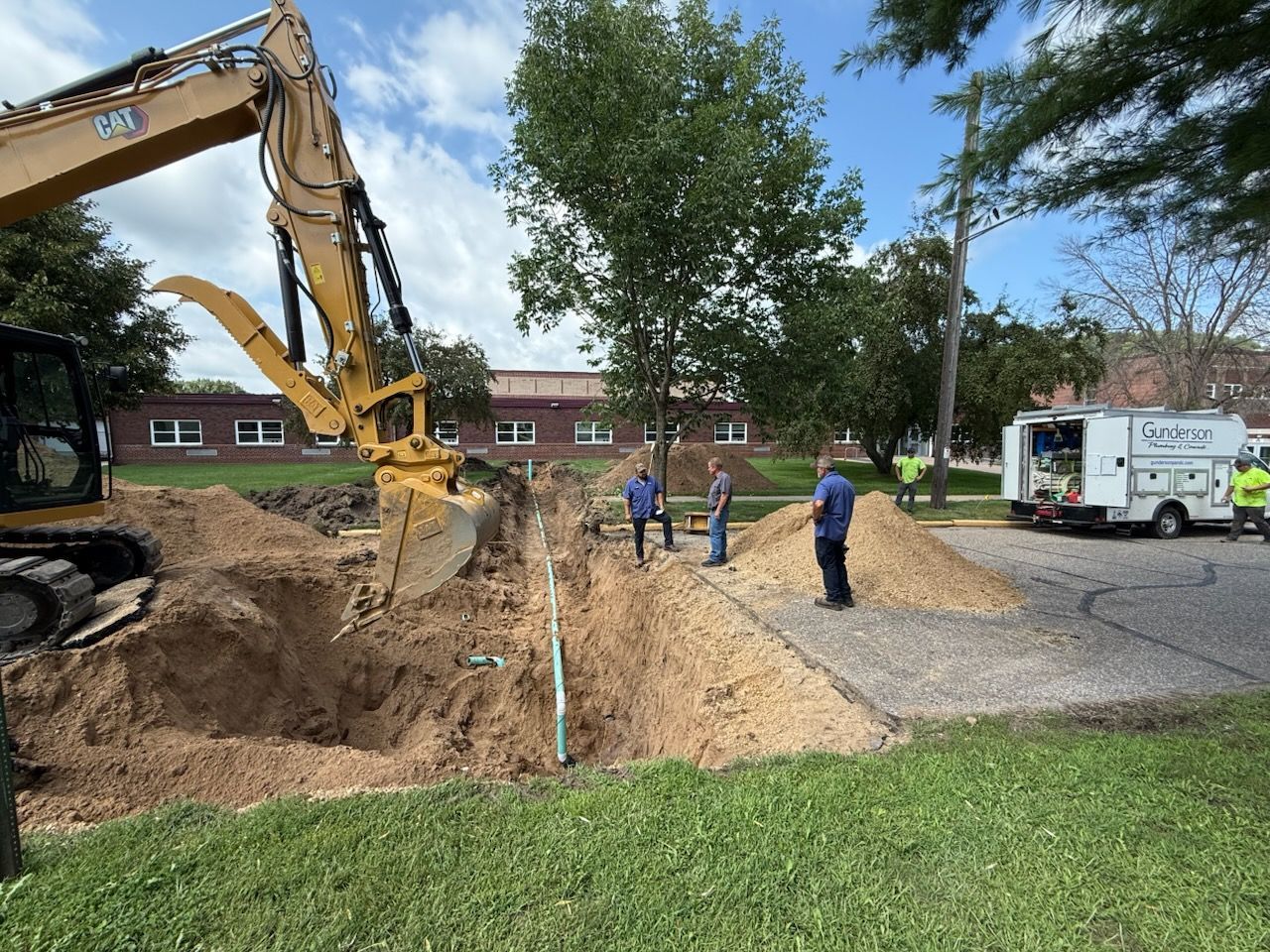 An excavator digs a trench on grass, with workers standing nearby. A service van is parked on the street.