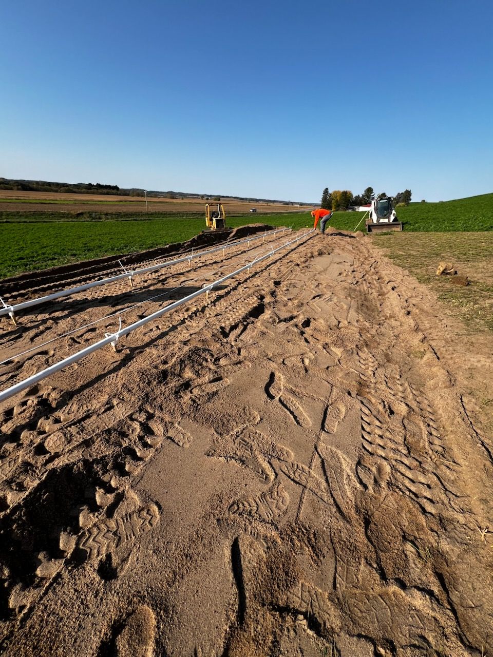 Construction site in field; machinery, pipelines, dirt, and green vegetation under a clear blue sky.