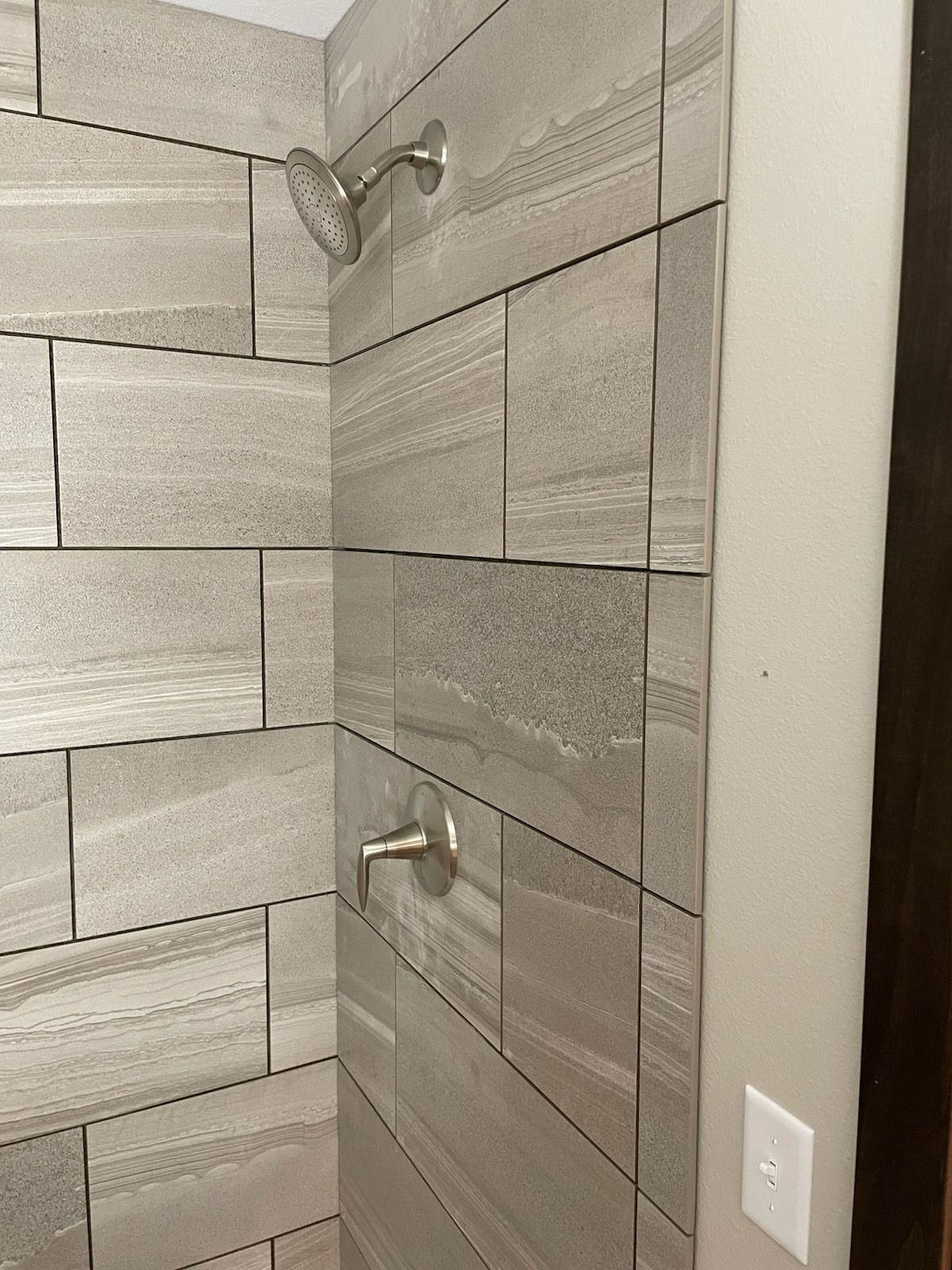 Shower with gray tiled walls, silver showerhead and faucet, and light beige wall with electrical outlet.