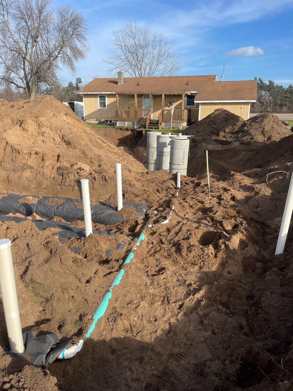 Septic system installation in progress. Brown soil surrounds pipes and concrete tanks near a house.