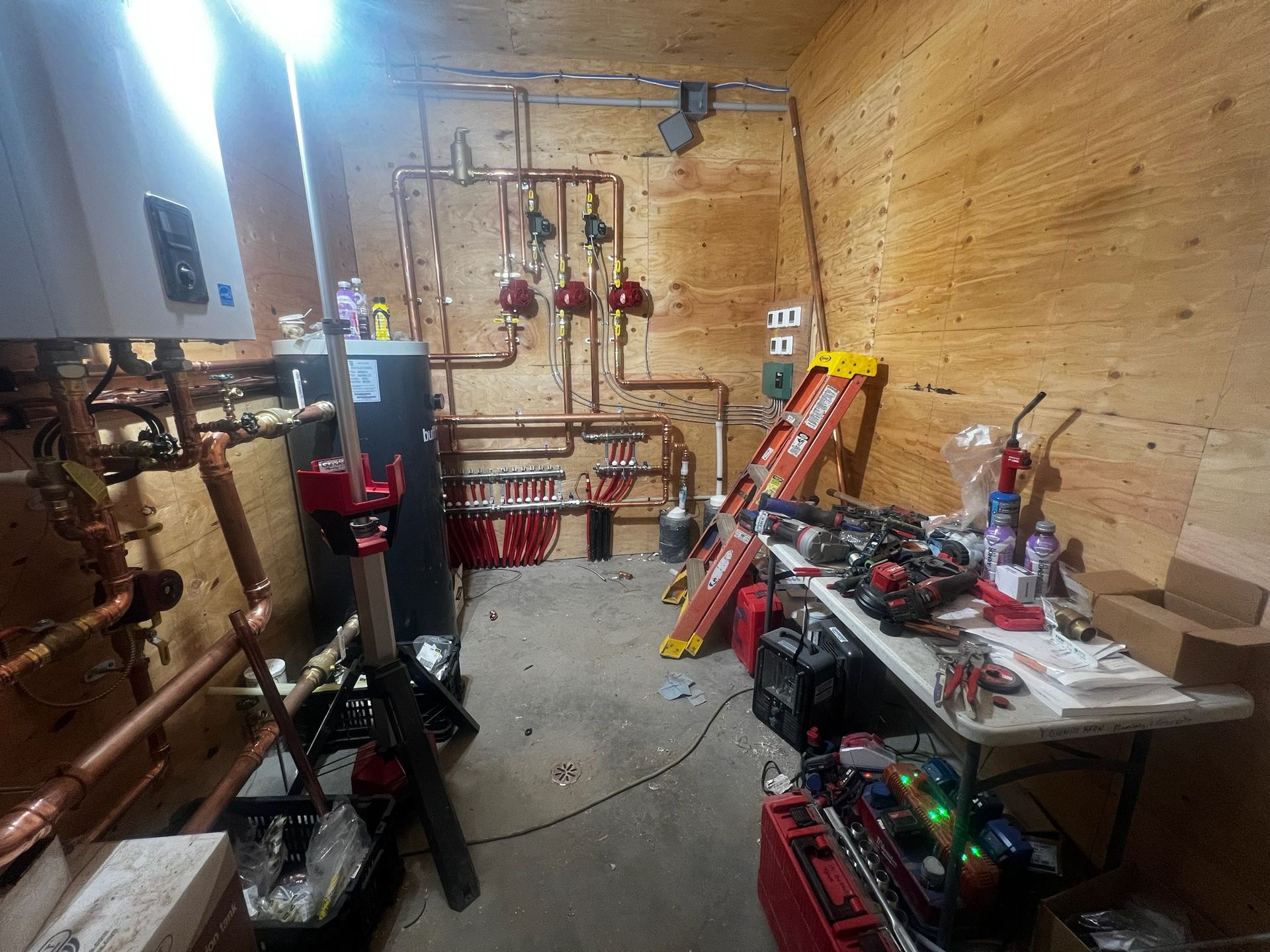 A cluttered utility room with copper pipes, equipment, tools, and a ladder.