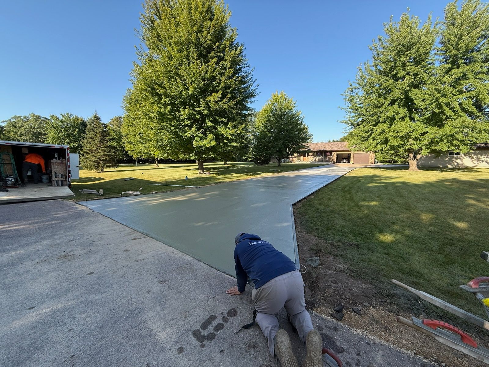Person working on a concrete driveway under a blue sky, near trees and a grassy lawn.