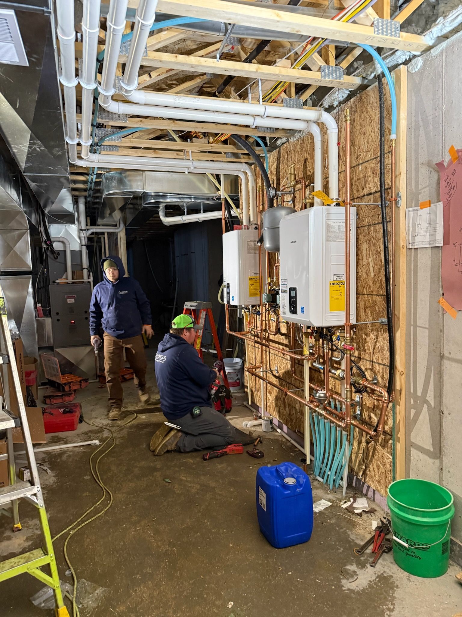 Two workers installing pipes and equipment in a room. White pipes and water heaters on a wood-paneled wall.
