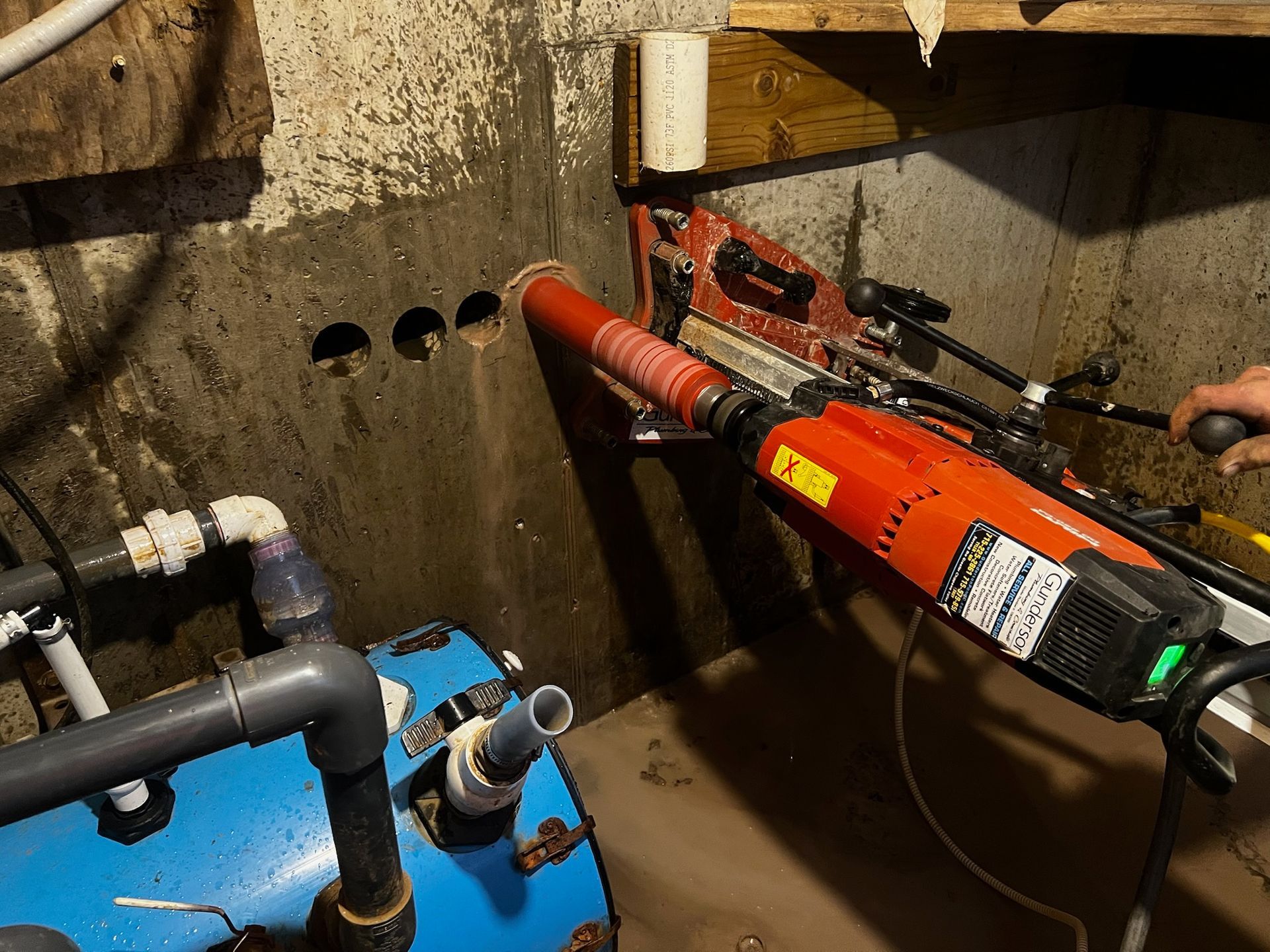 Red coring drill making holes in a concrete wall, near a blue water tank.