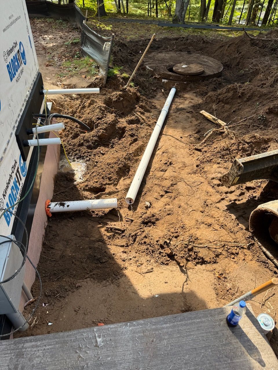 Construction site with white pipes, dirt, and a small excavator near a building.