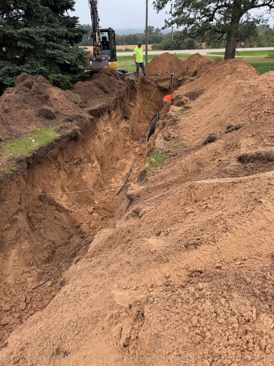 Man in trench digging a ditch for utilities; heavy equipment nearby. Brown soil.