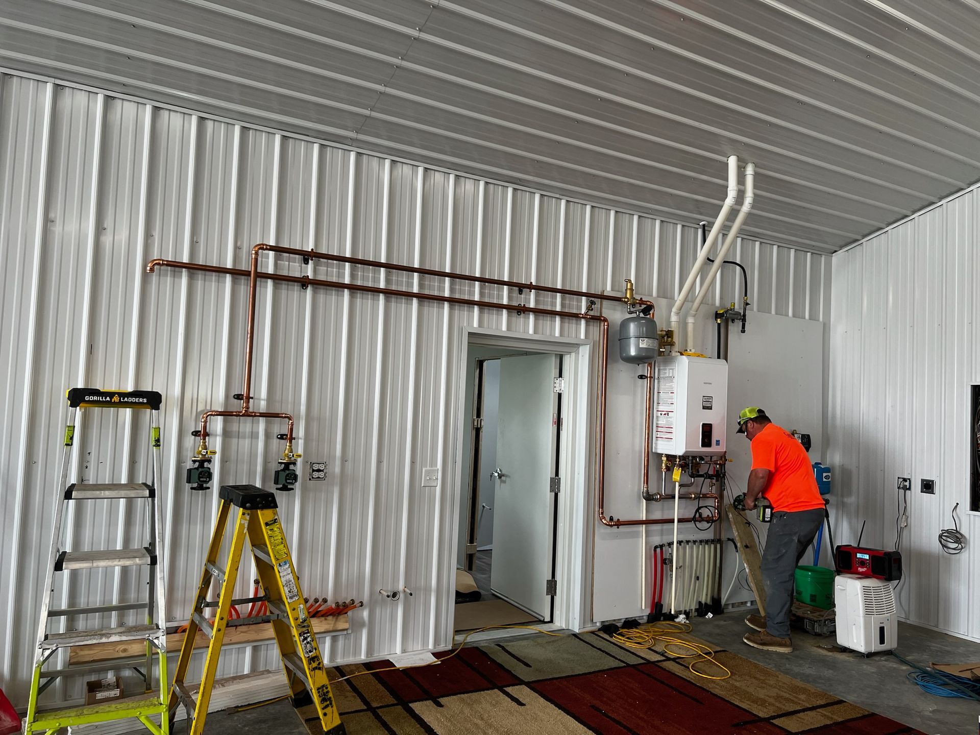 A worker installs plumbing in a room with white corrugated walls and ceiling. Copper pipes are visible.