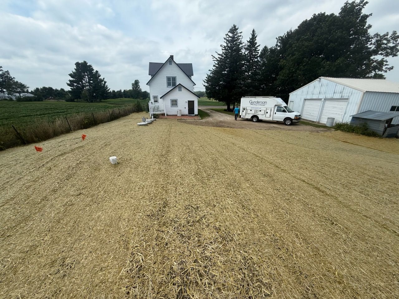 A field of oats leads to a white farmhouse and a van parked next to a barn on a cloudy day.