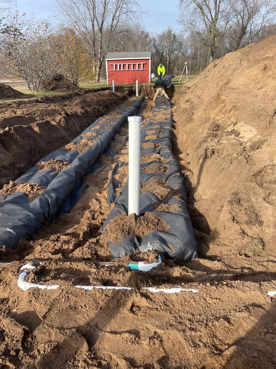 Installation of a new septic system with two wrapped trenches, white pipes, and a worker in a red building in the background.
