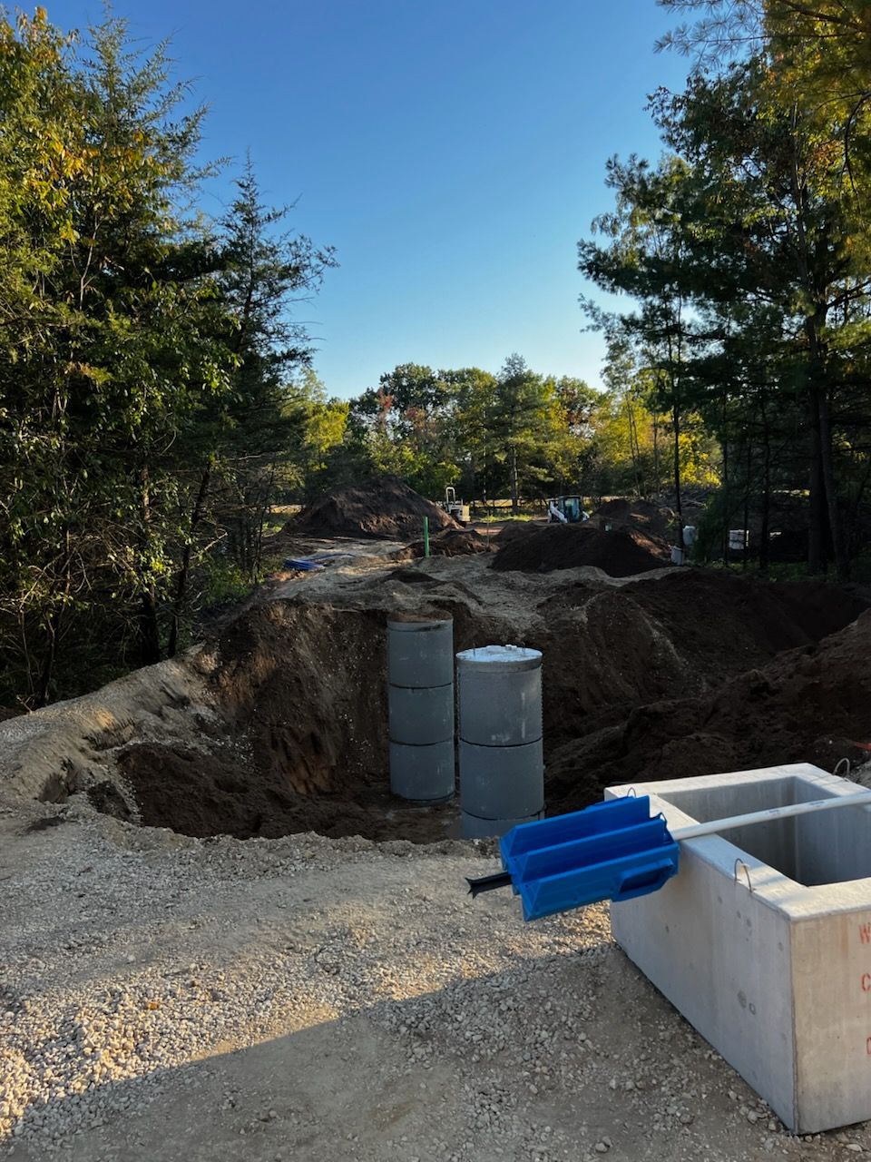 Construction site with concrete tanks and blue tool, surrounded by dirt and trees under blue sky.