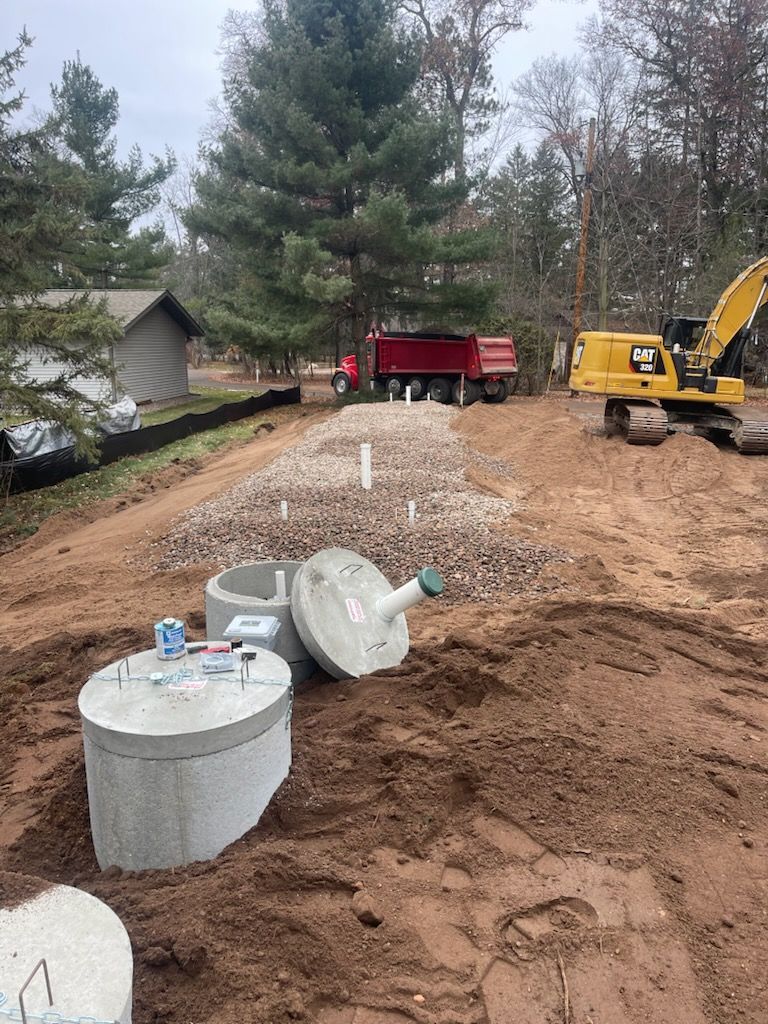 Construction site with septic tanks and a backhoe, in front of a dump truck and treeline.