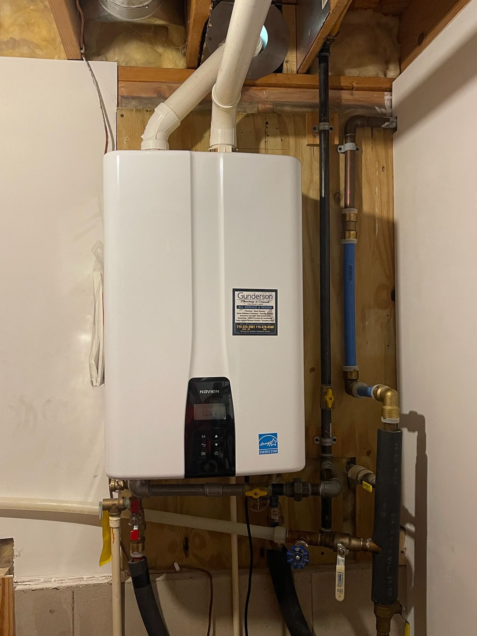 White tankless water heater on a wooden wall with pipes, vents, and a control panel.
