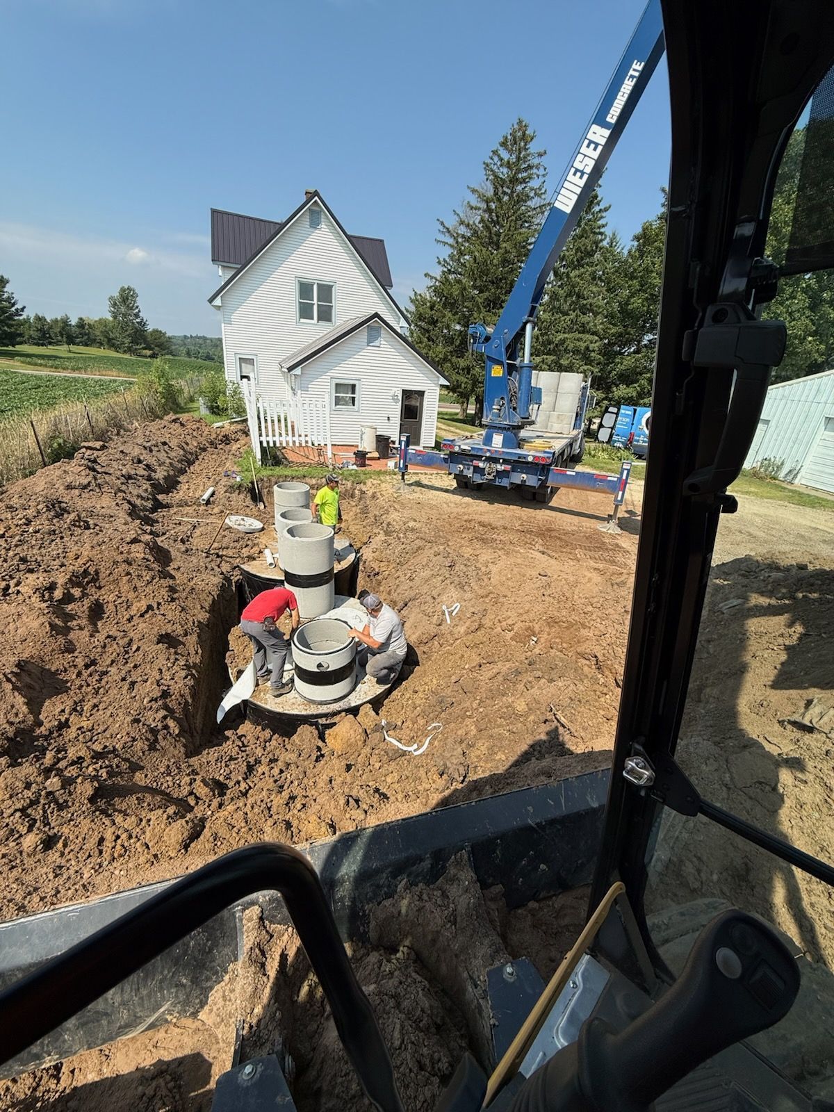 Construction site: Workers installing concrete rings near a house. A blue crane is visible, along with an excavator cab.