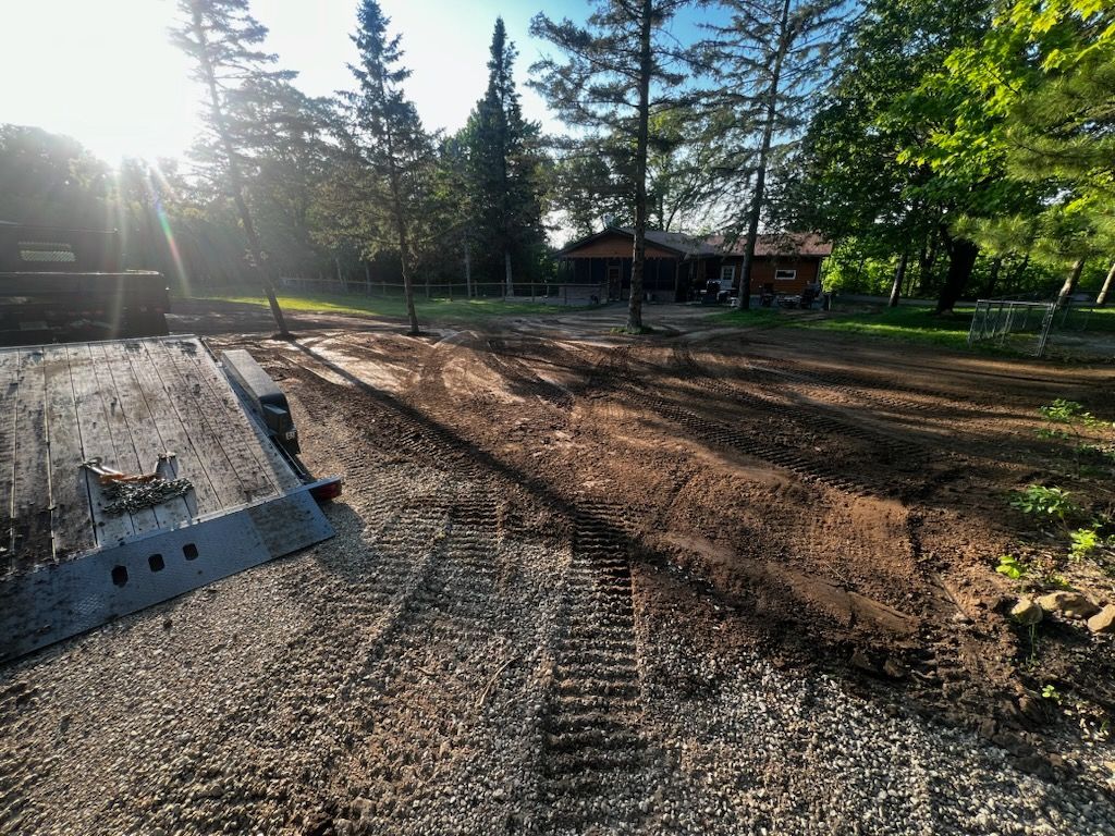 Dirt work in front of a house, with a trailer and gravel driveway; trees and sun in the background.