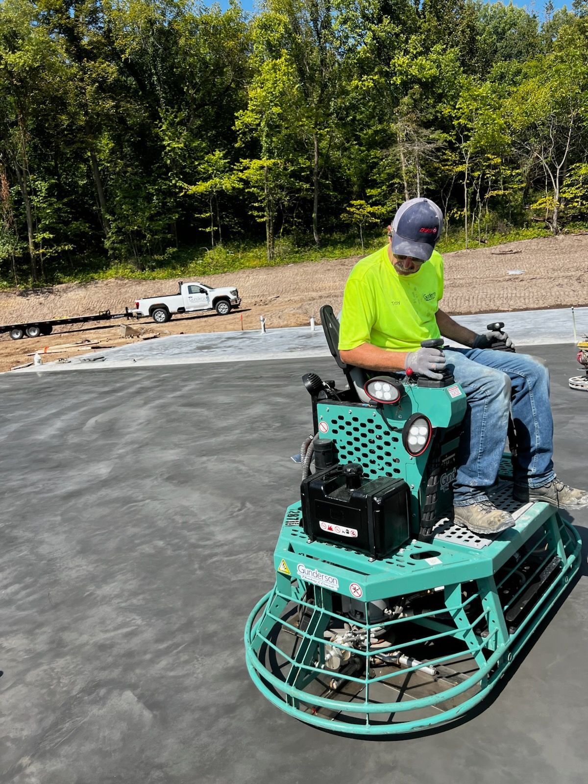 Man operating a power trowel on a concrete surface, outdoors. Wearing a neon shirt and seated, truck in the background.